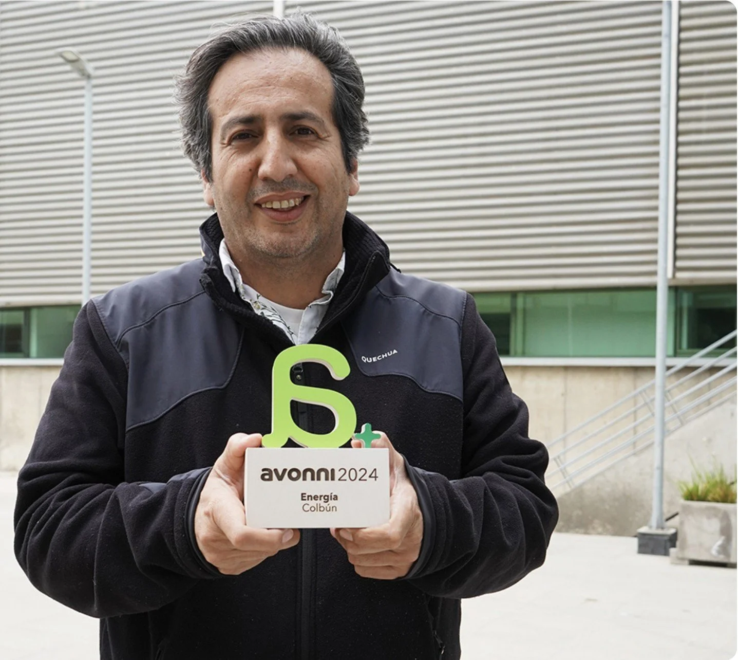 A man holding an award for Avonni 2024 in energy, outdoor setting with modern building in background.