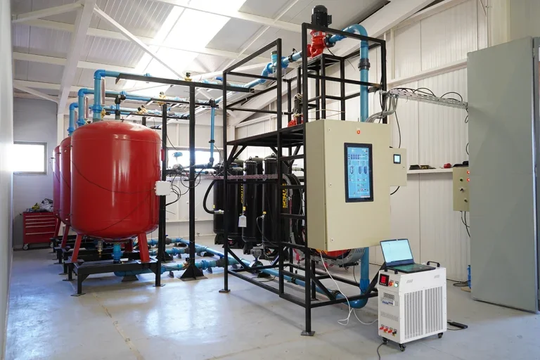 Industrial water treatment system with a large red pressure tank, blue pipes, black filtration units, control panel, and cooling unit inside a white-walled facility.