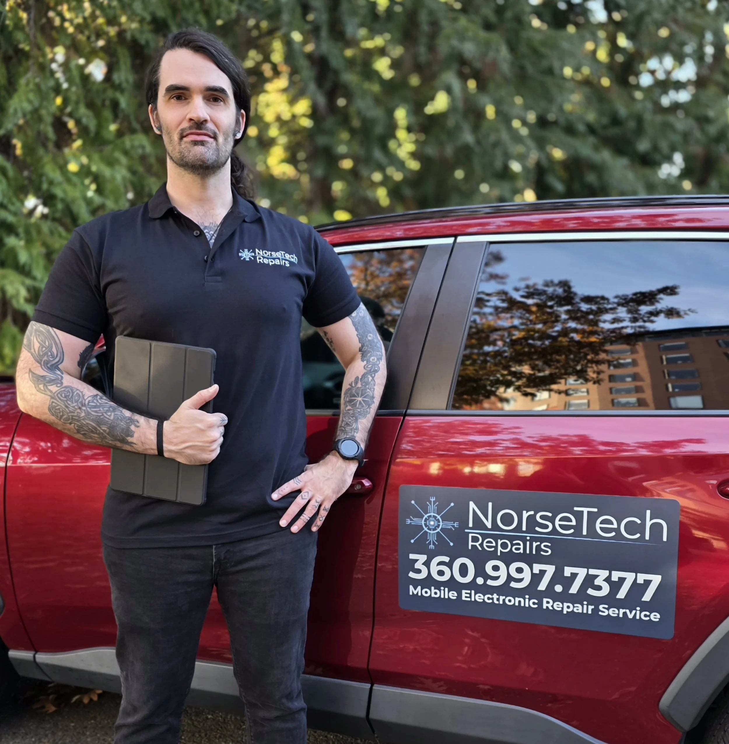 Tattooed mobile electronics repair technician holding a tablet beside a red NorseTech Repairs service vehicle.