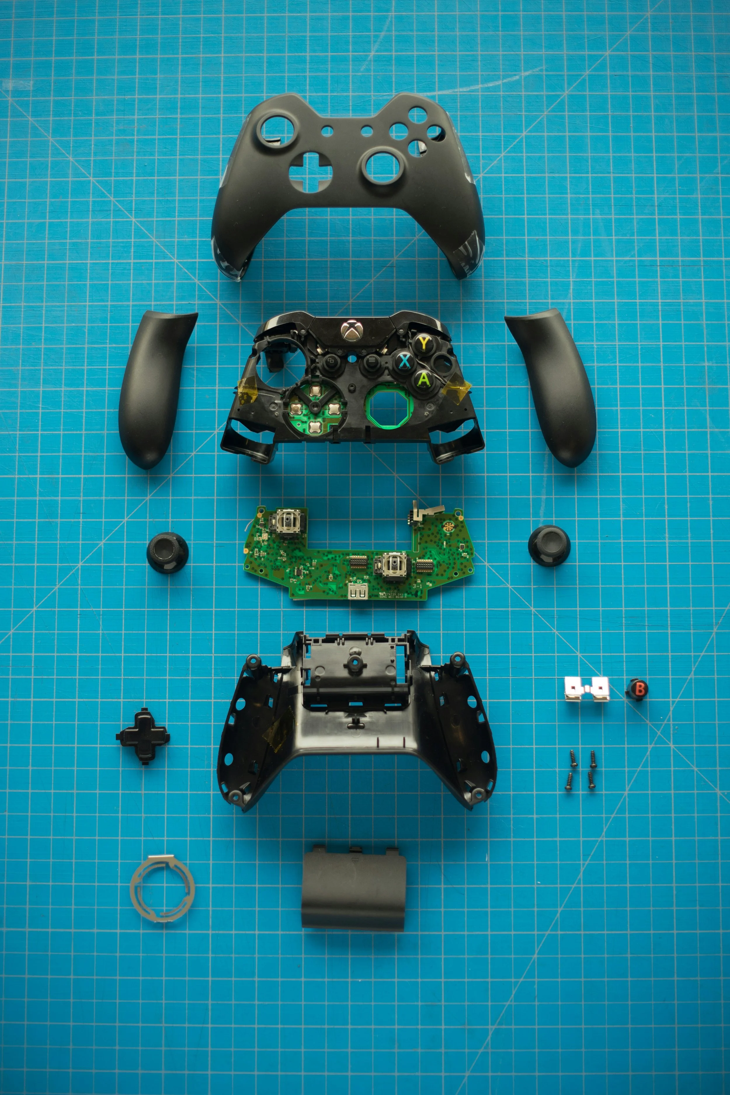 Disassembled Xbox game controller with shell, circuit board, buttons, thumbsticks, grips, and screws arranged on a blue cutting mat.