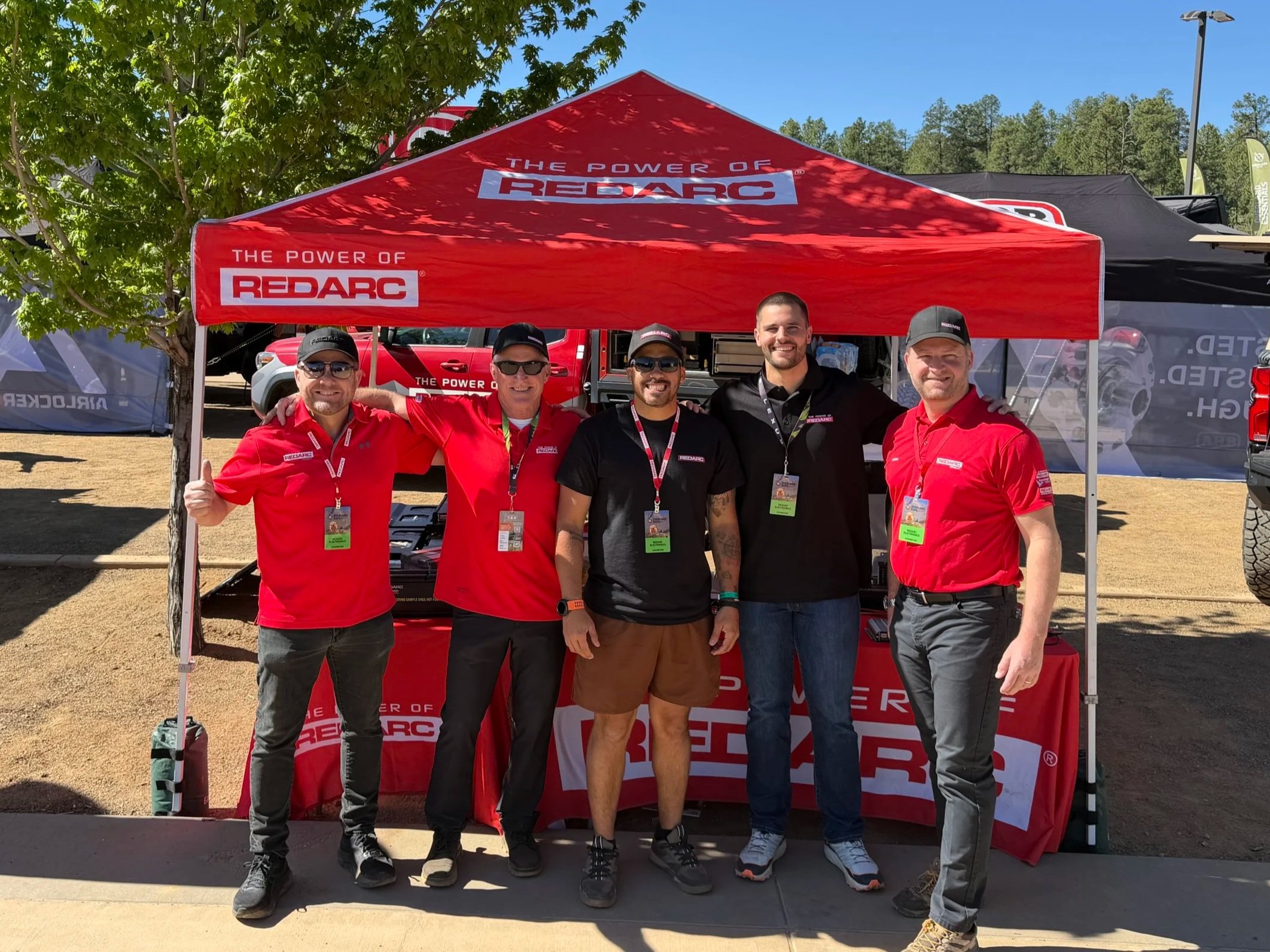 A group of five men standing under a red tent with "Redarc" branding, outdoors on a sunny day. They are smiling, wearing casual and branded clothing, and some have sunglasses and hats. Behind them are trees, a blue sky, and several tents or booths.