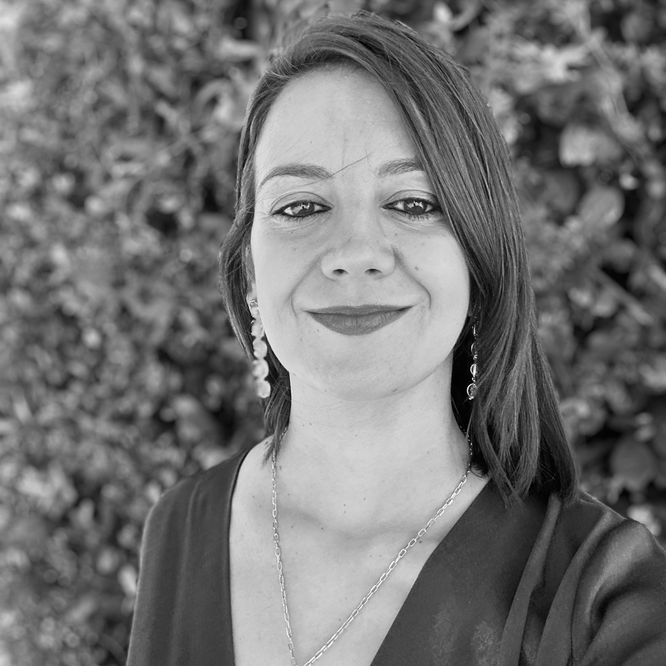 Retrato de una mujer con cabello largo y peinado a un lado, usando pendientes y cadenas, con fondo de plantas difusas.