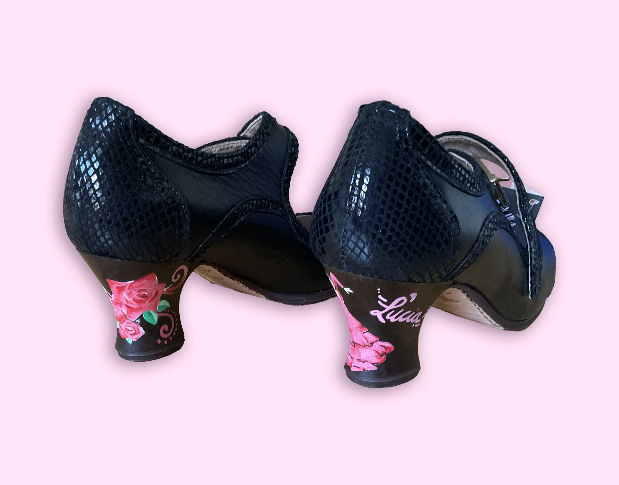 Zapatos de tacón negros con textura de malla y adornos florales rosas y verdes, decorados con etiquetas colgadas.