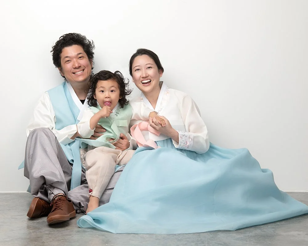 A happy family of three, parents and a young boy sitting on the floor and smiling at camera, with the parents wearing traditional Korean clothing, known as hanbok.
