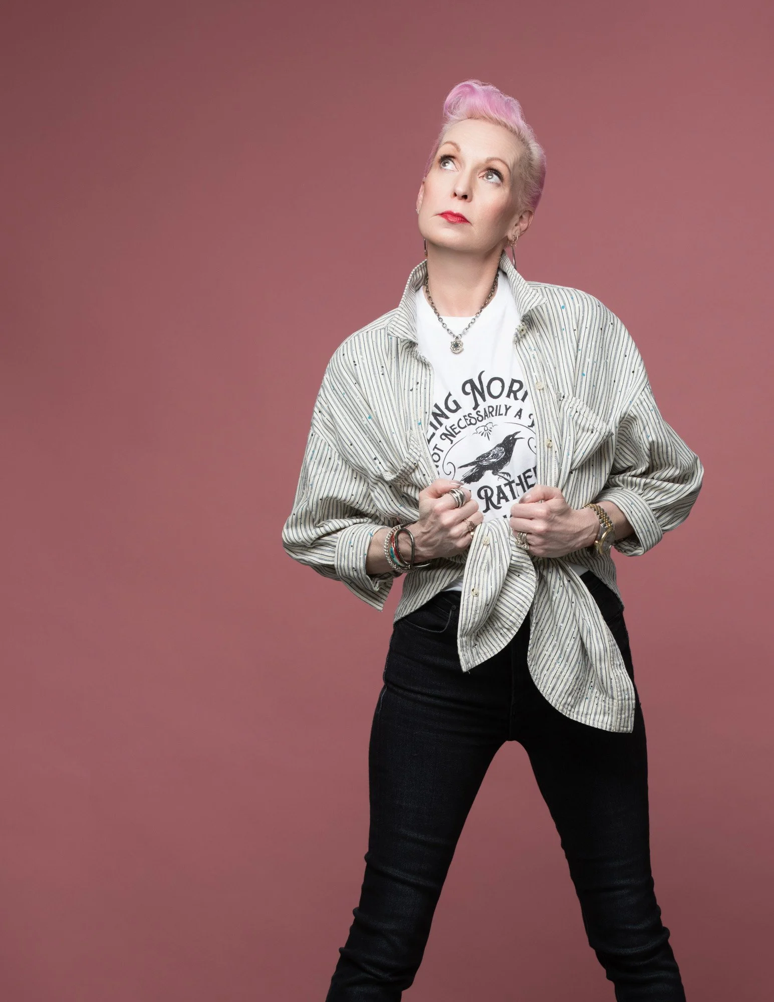 A woman with pink hair in a vintage pompadour hairstyle, wearing a graphic T-shirt with a crow and text, layered with a striped shirt tied at the waist, and black jeans, standing against a plain mauve background, looking upward.