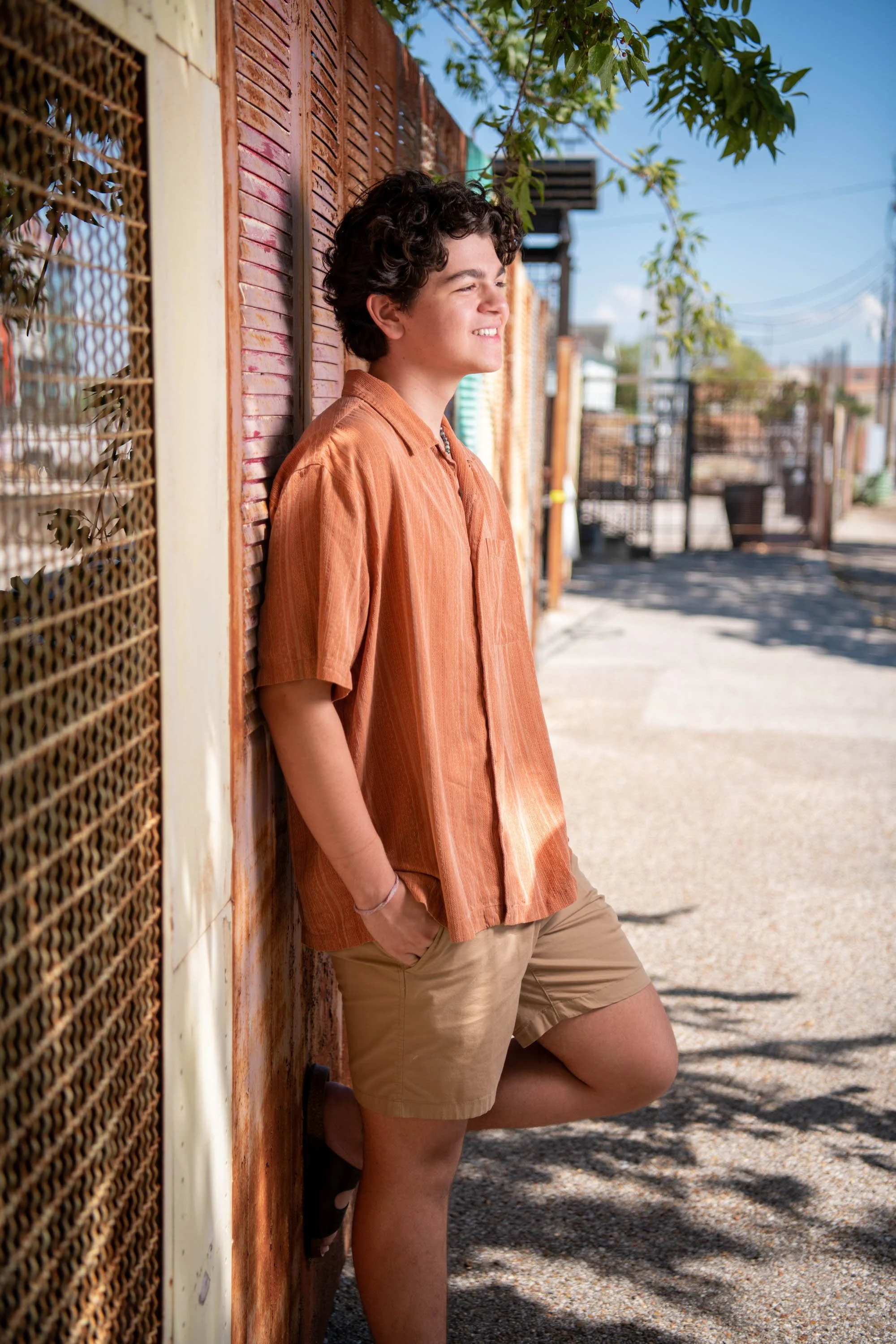 A young person with curly dark hair, wearing an orange short-sleeved shirt and beige shorts, leans against a rusted metal fence with a smile, standing outside on a sunny day with trees and a clear blue sky in the background.