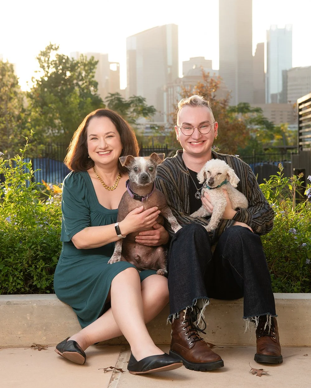 Two people sitting outdoors at sunset in a city park with two small dogs, green plants, and tall city buildings in the background, smiling at the camera.