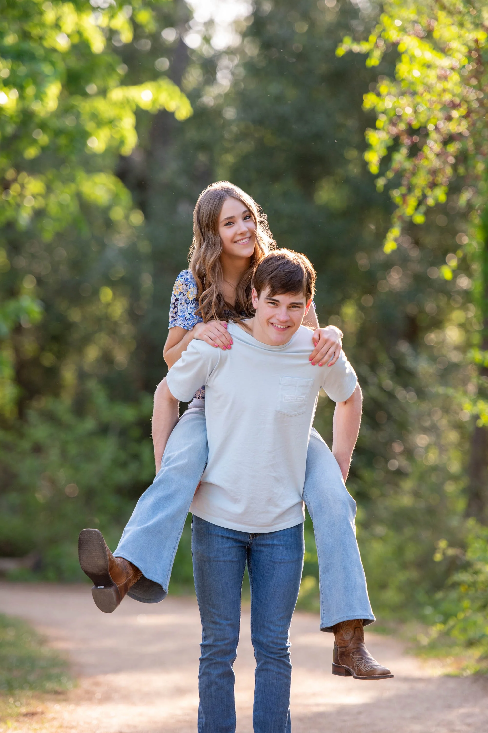 A young man giving a piggyback ride to a young woman in a park during daytime, both smiling and enjoying the outdoors with green trees in the background.