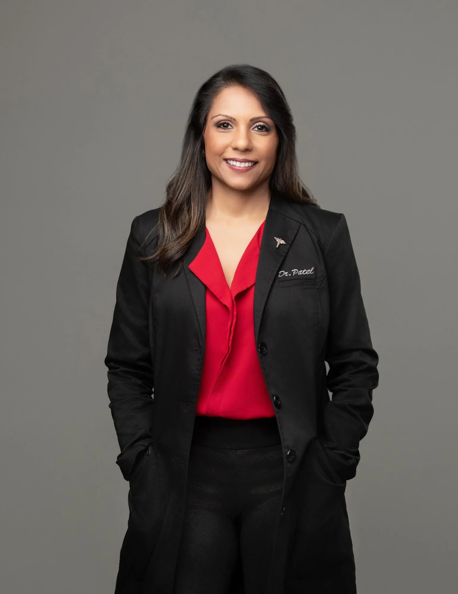 A woman smiling, wearing a black blazer and a red blouse, standing against a plain grey background.