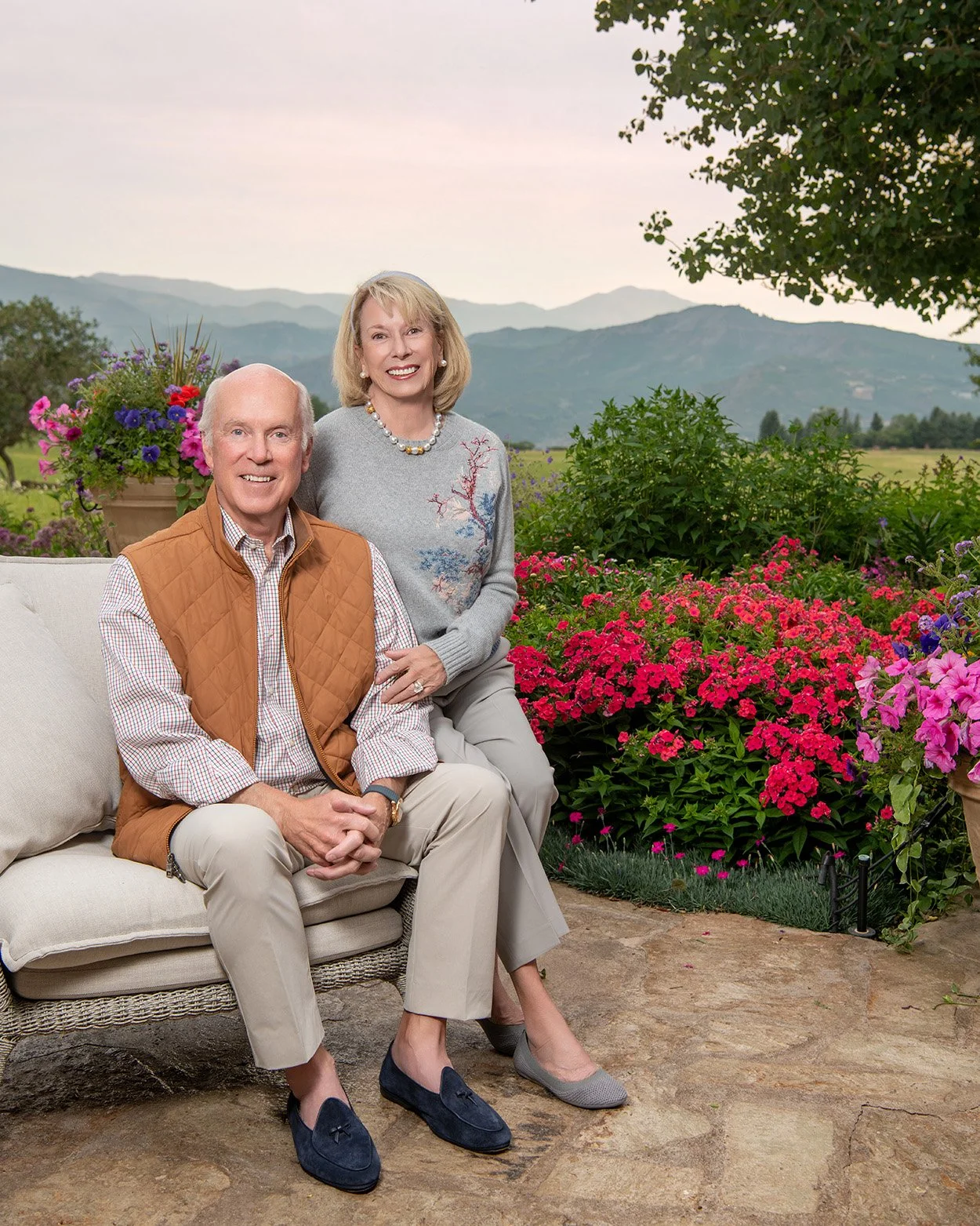 An older man and woman smiling outdoors on a patio surrounded by colorful flowers with mountains in the background.