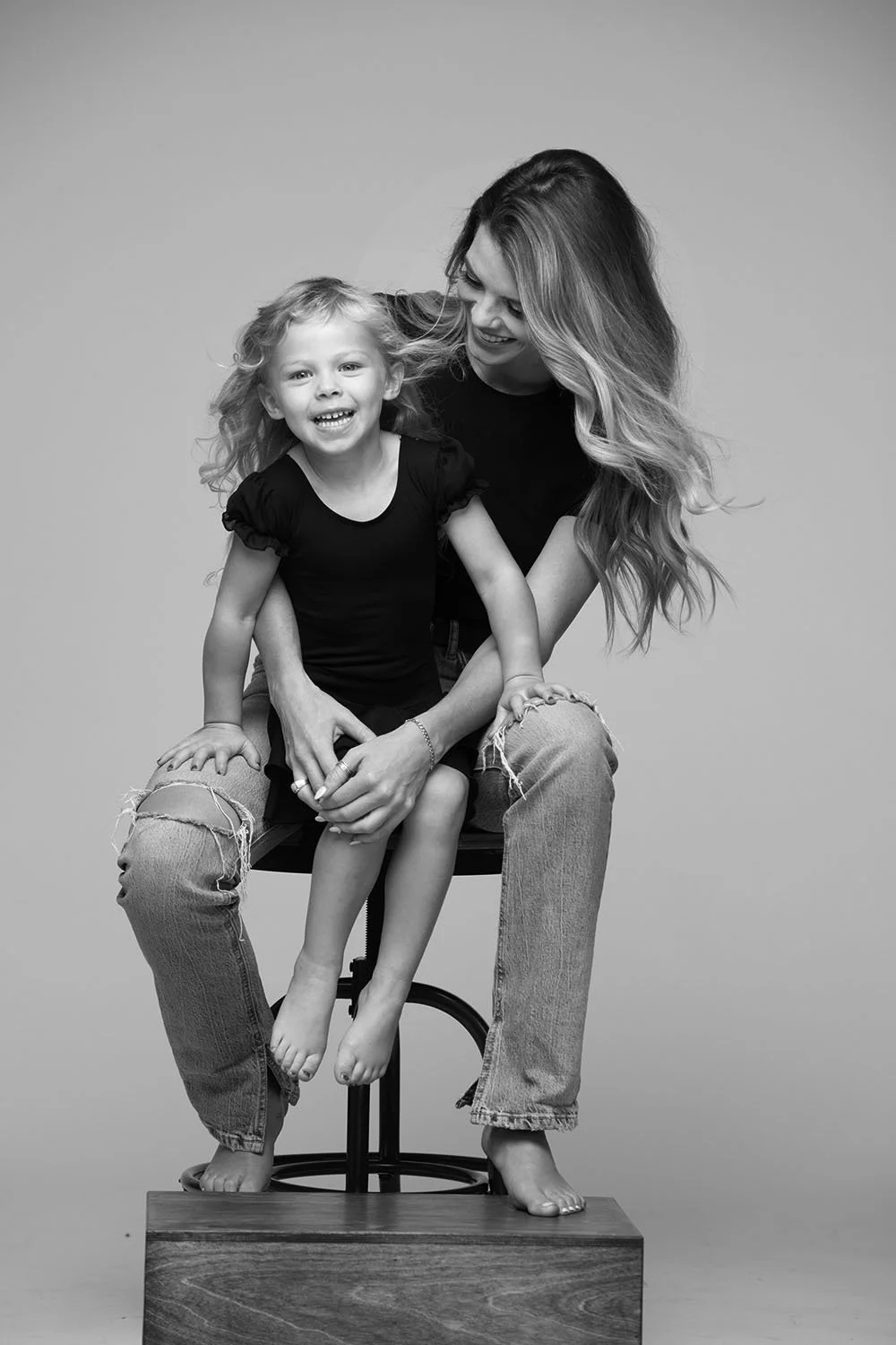 Black and white photo of a woman and a young girl smiling and playing together. The woman has long, wavy hair and is sitting on a stool, wearing ripped jeans and a black shirt. The girl is sitting on the woman's lap, also smiling, with curly hair, wearing a black dress, and both are barefoot.