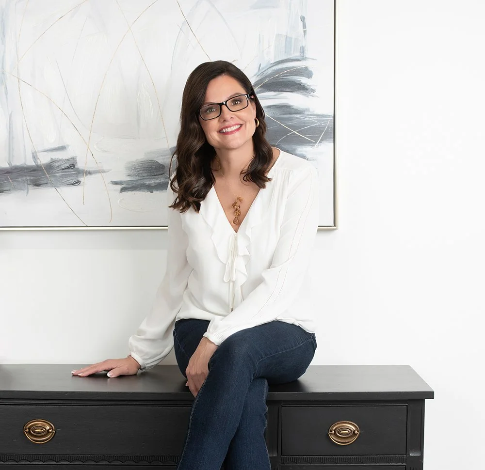 A woman with glasses, shoulder-length dark hair, and a white blouse, sitting on a black cabinet in front of an abstract painting, smiling at the camera.