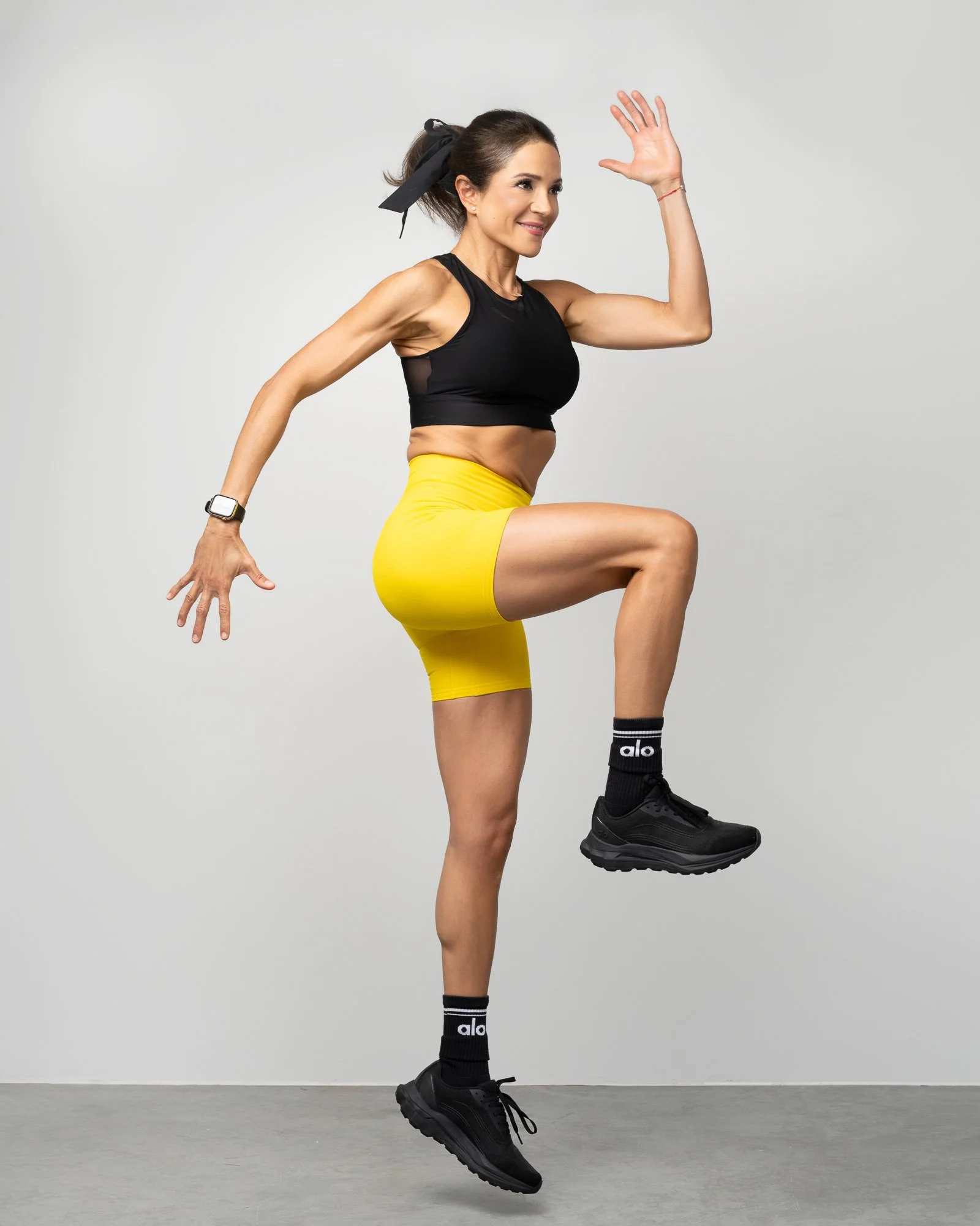 A woman with dark hair tied with a black ribbon, wearing a black sports bra, yellow shorts, and black sports shoes, is balancing on one leg with her right knee raised, left hand on her knee, and right hand raised in a waving gesture, against a plain 