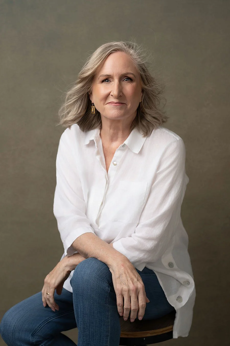 An older woman with shoulder-length gray hair and light skin, sitting on a wooden stool against a neutral background. She is wearing a white button-up shirt and blue jeans, looking directly at the camera with a slight smile.