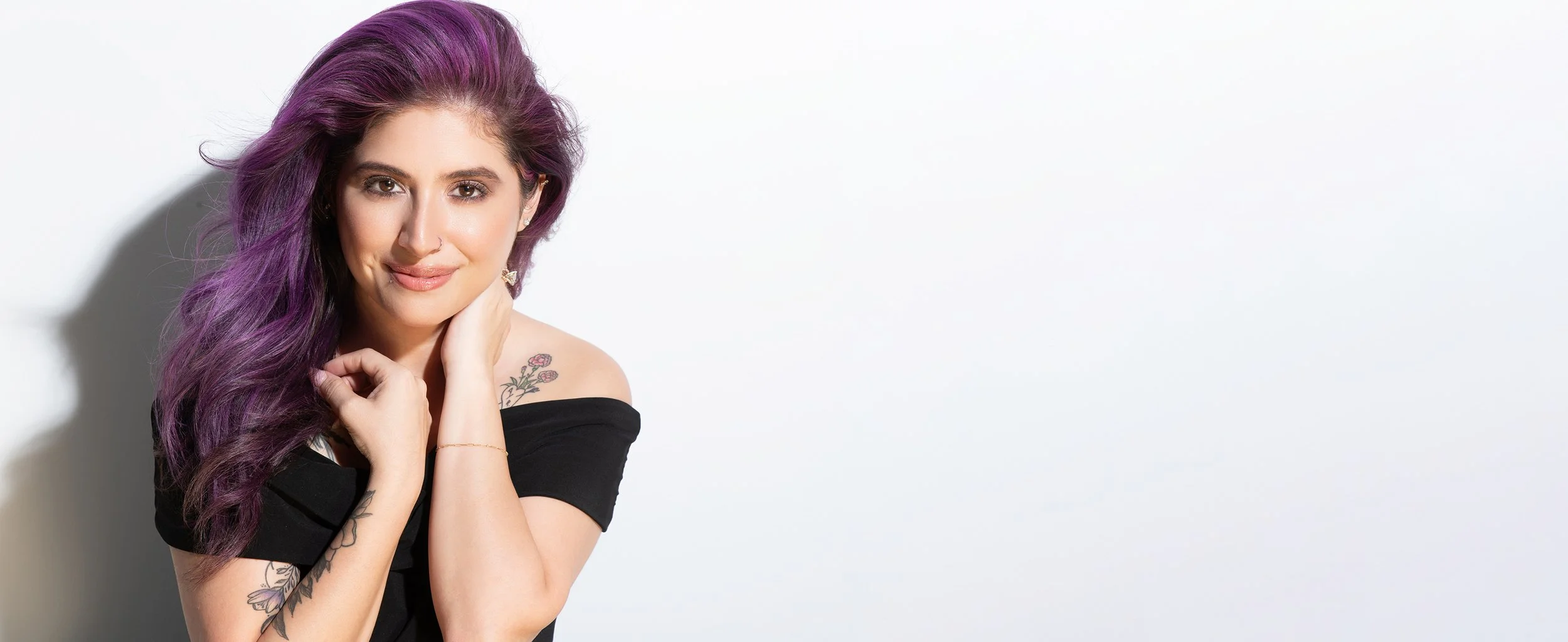 A young woman with purple hair, wearing a black off-the-shoulder top, smiling and posing with her hand near her face against a white background.
