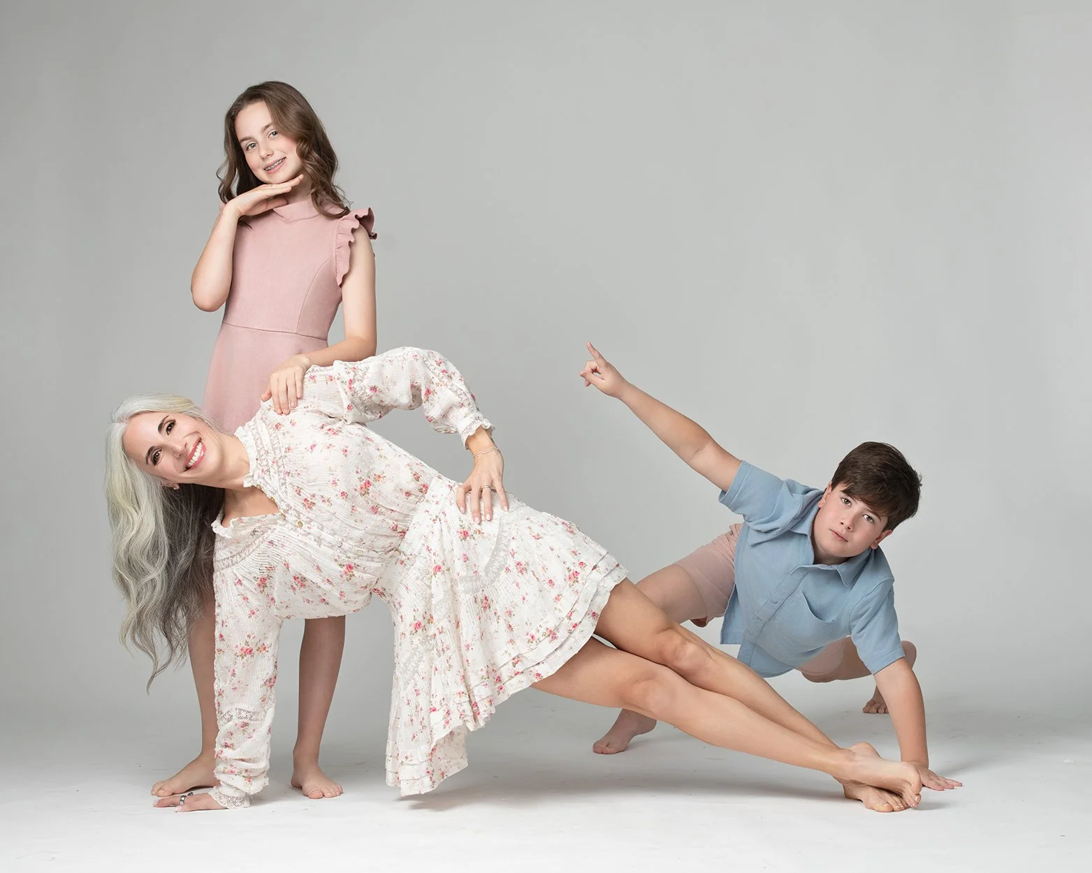 A woman with long gray hair in a white floral dress performs a yoga pose on a plain white background. A young girl in a pink dress stands behind her, smiling and touching her chin, while a young boy in a blue shirt and shorts leans on the woman's bac