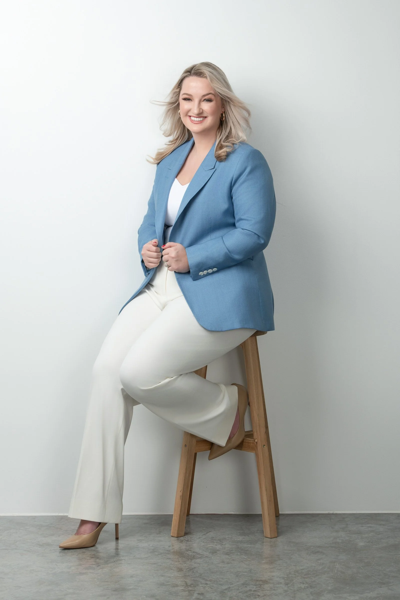 A woman with blonde hair, wearing a blue blazer, white top, and white pants, is sitting on a wooden stool against a plain white wall, smiling and winking.