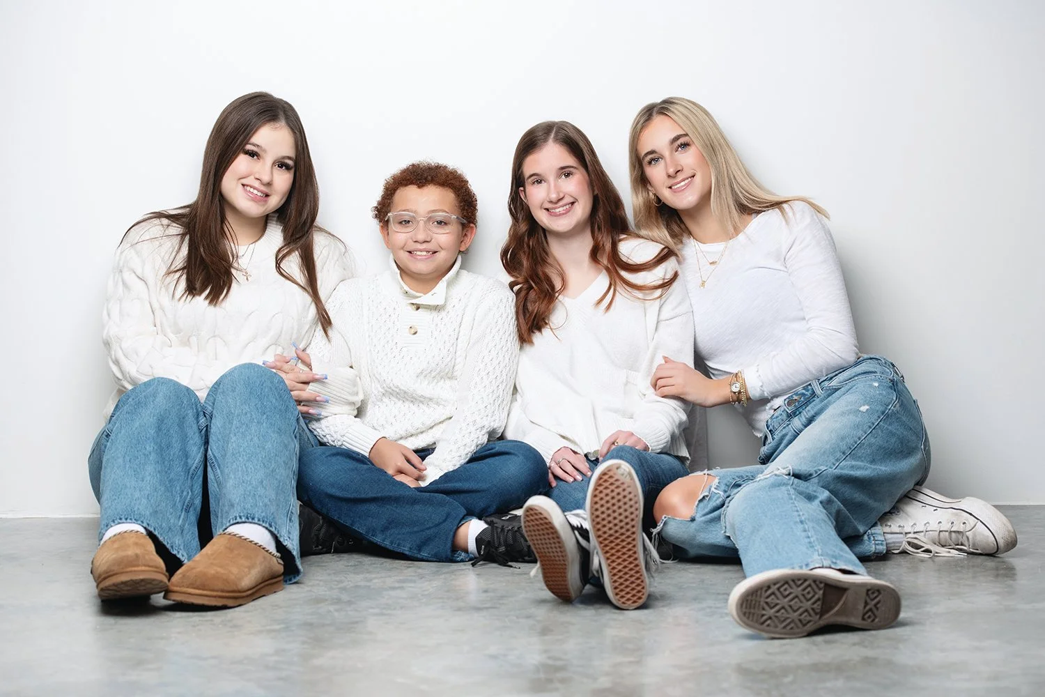 Four kids sitting on the floor against a white background, smiling at the camera. They are dressed casually in sweaters and jeans.