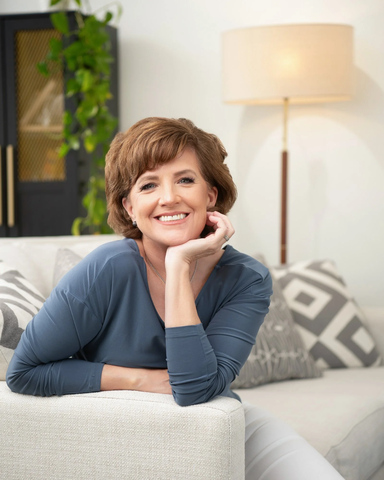 Portrait of a smiling woman with short brown hair, wearing a blue blouse, resting her chin on her hand, sitting on a beige couch with patterned cushions in a well-lit modern living room.