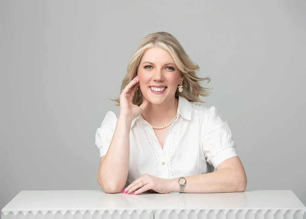 Portrait of a smiling woman with blonde hair, wearing a white blouse, pearl necklace, earrings, and a wristwatch, sitting at a white table against a gray background.