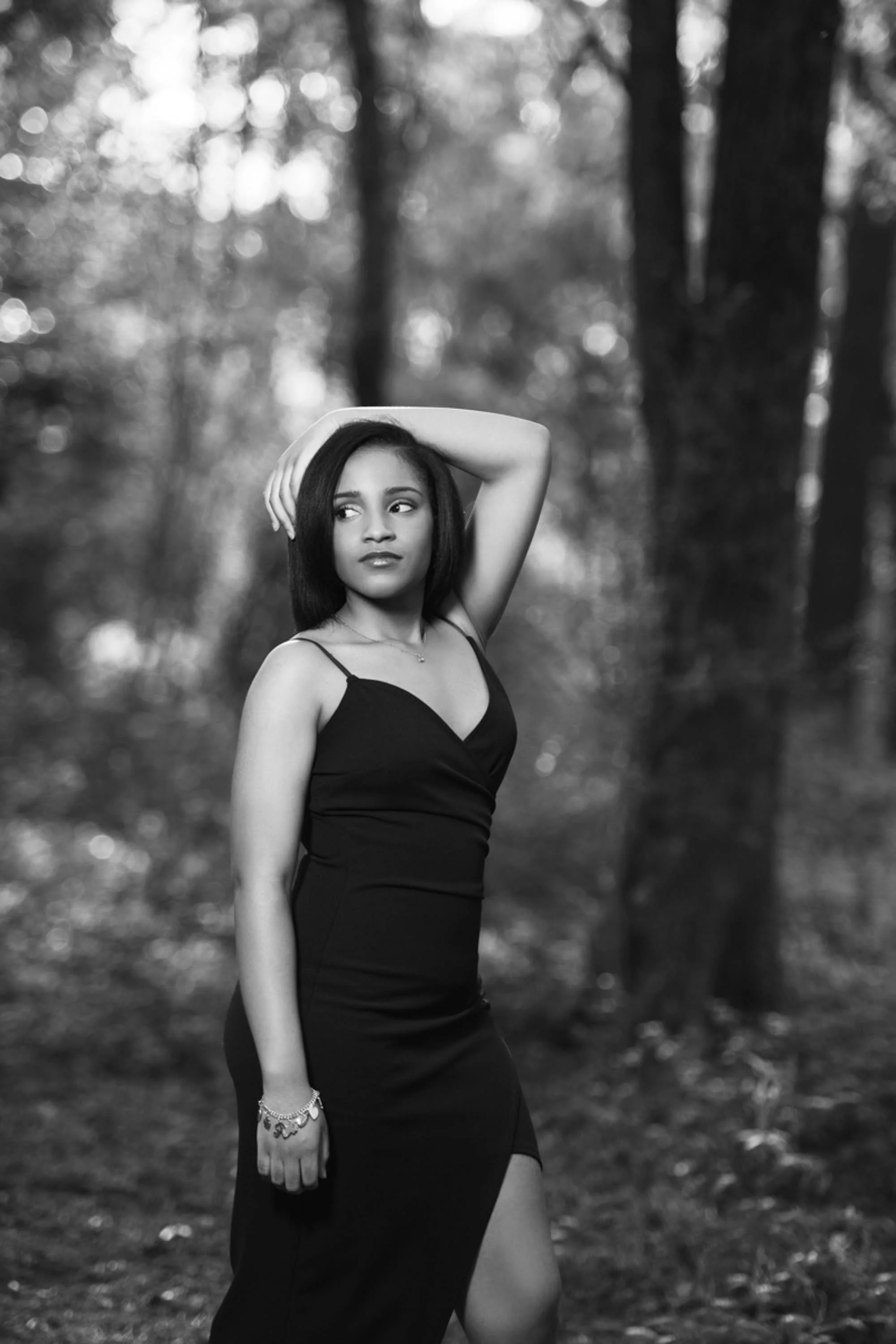 A young woman with dark, straight hair wearing a black dress with thin straps, standing outdoors in a forest. Her right arm is raised with her hand resting on her head, and she is looking to her left. The background is blurred with trees and light fi