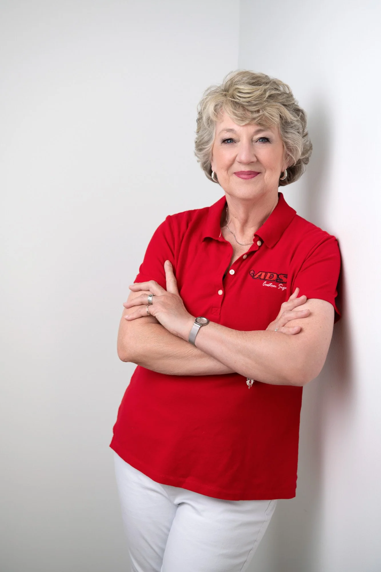 A woman with blonde curly hair wearing a red polo shirt and white pants, standing against a white background with arms crossed, smiling.