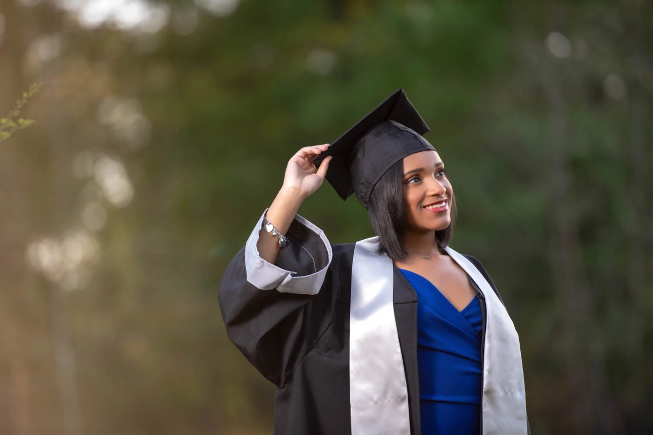A young woman in a graduation gown and cap adjusting her cap outdoors, smiling with a blurred green background.