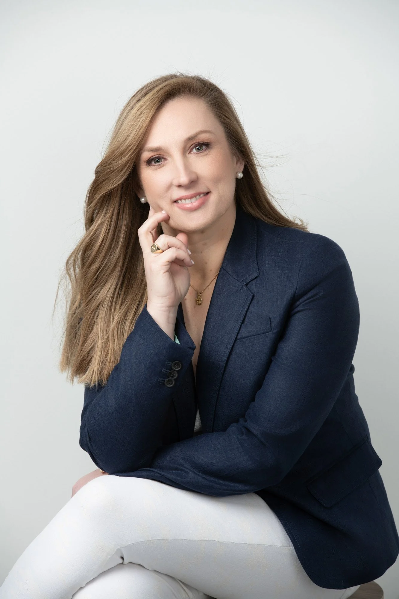 A woman with long, wavy light brown hair poses against a plain gray background. She is wearing a navy blazer, pearl earrings, a gold ring, and a gold necklace with a pendant. She is sitting with her left arm resting on her thigh and her right hand to