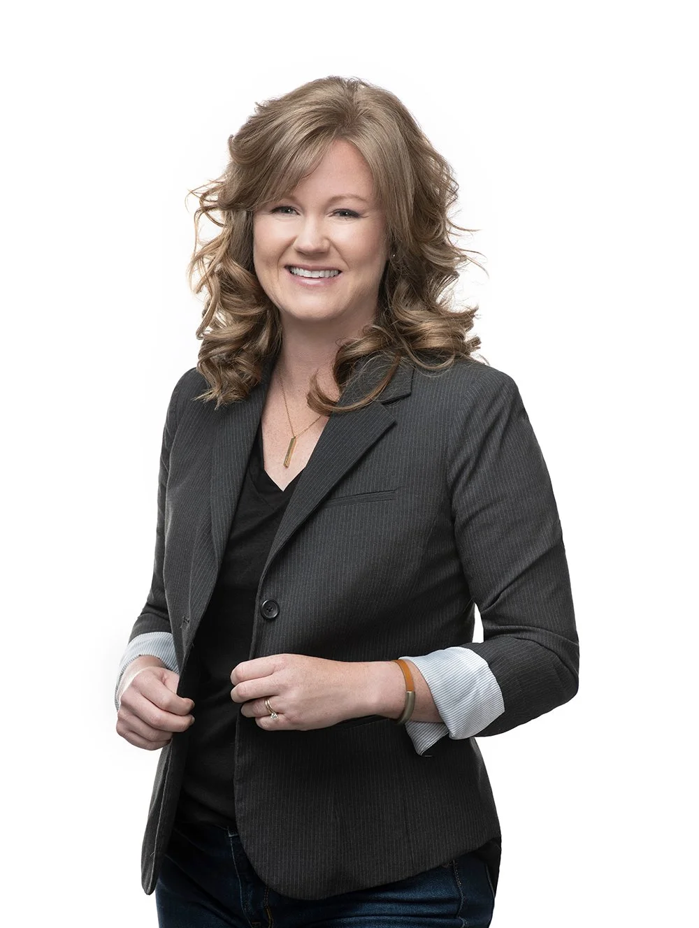 A smiling woman with red hair in a black blazer, wearing a necklace and a bracelet, standing against a white background.