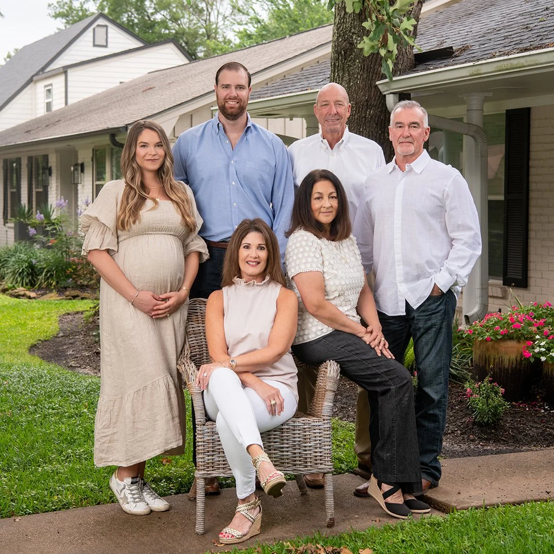A group of six people, mixed genders and ages, posing outdoors in front of a house with a garden and trees, smiling for a photo.
