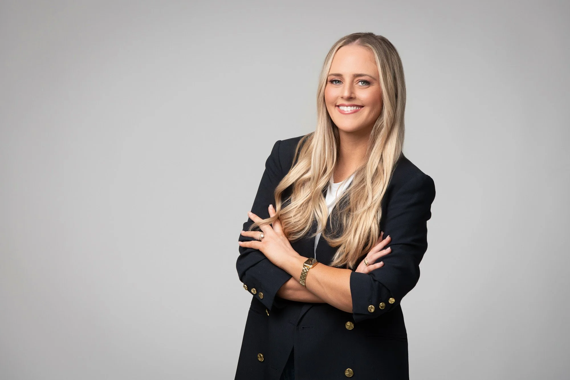A woman with long blonde hair, smiling and wearing a black blazer with gold buttons, stands with arms crossed against a light gray background.