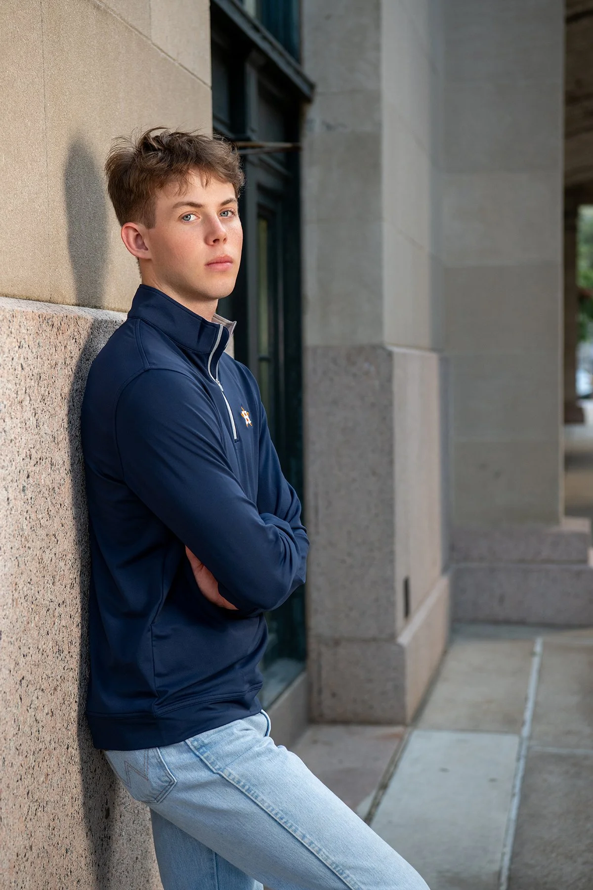 A young man with brown hair and fair skin leaning against a tan brick wall, wearing a navy blue zip-up jacket and light blue jeans, with arms crossed and looking at the camera.
