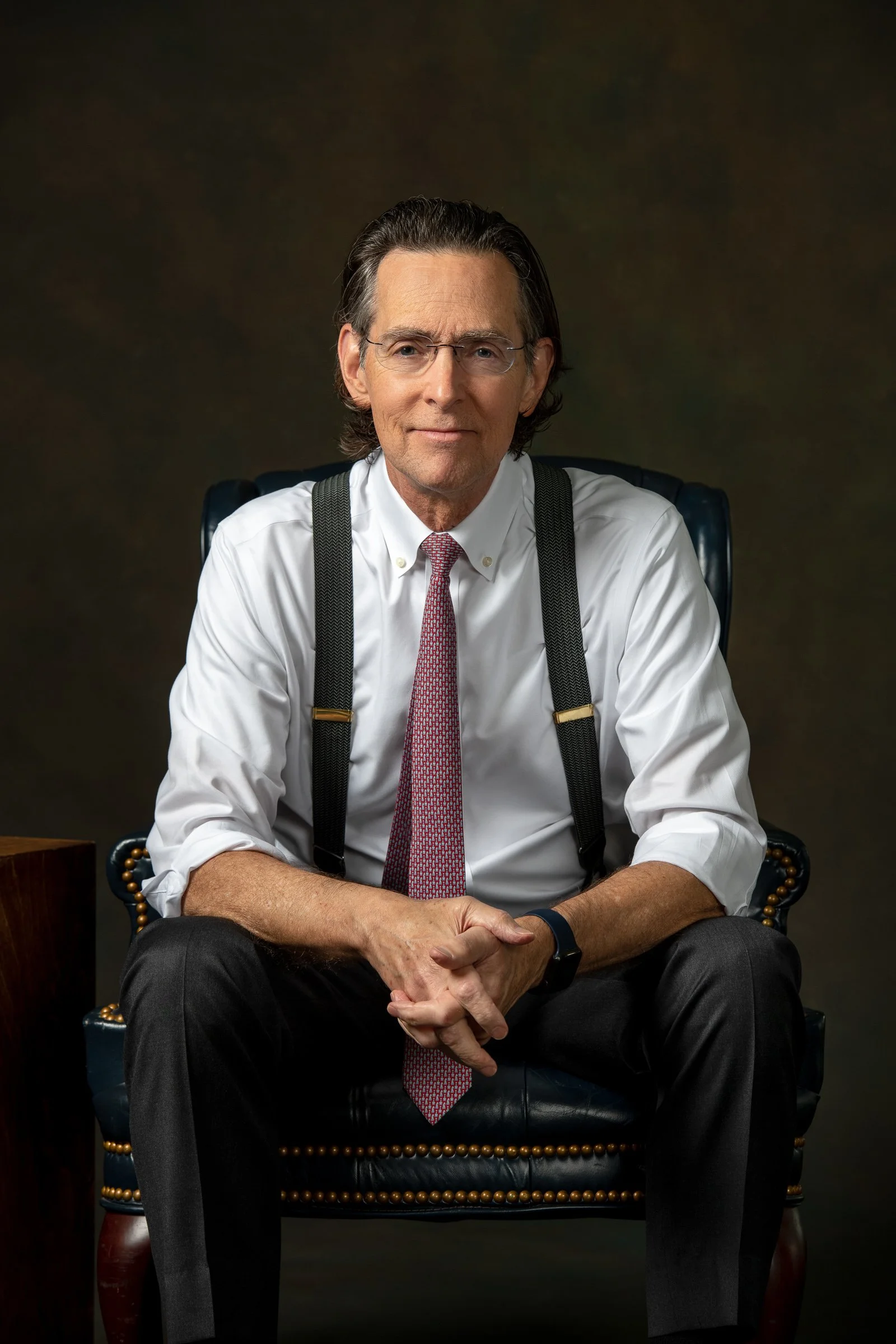 A middle-aged man with glasses, wearing a white dress shirt, red patterned tie, and suspenders, sitting in a black leather chair with brass nailhead trim, against a dark background.
