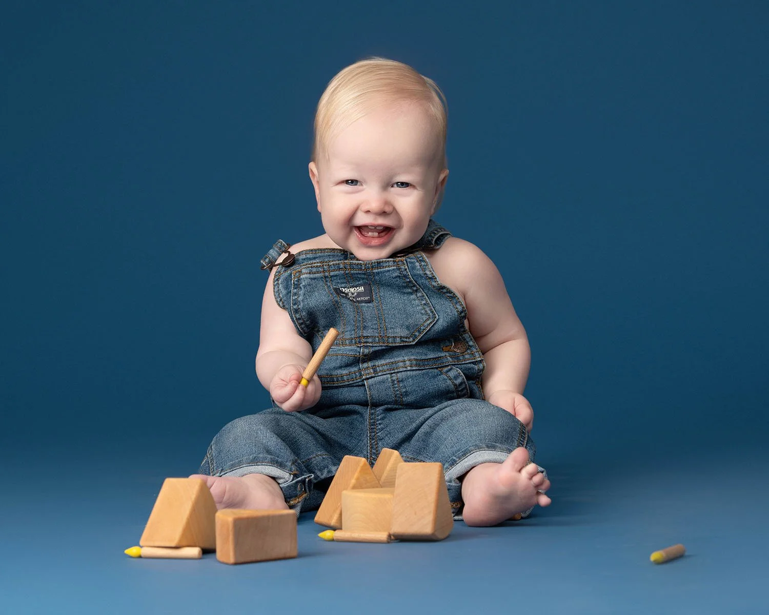 A smiling toddler in denim overalls playing with wooden blocks and a yellow marker on a blue background.