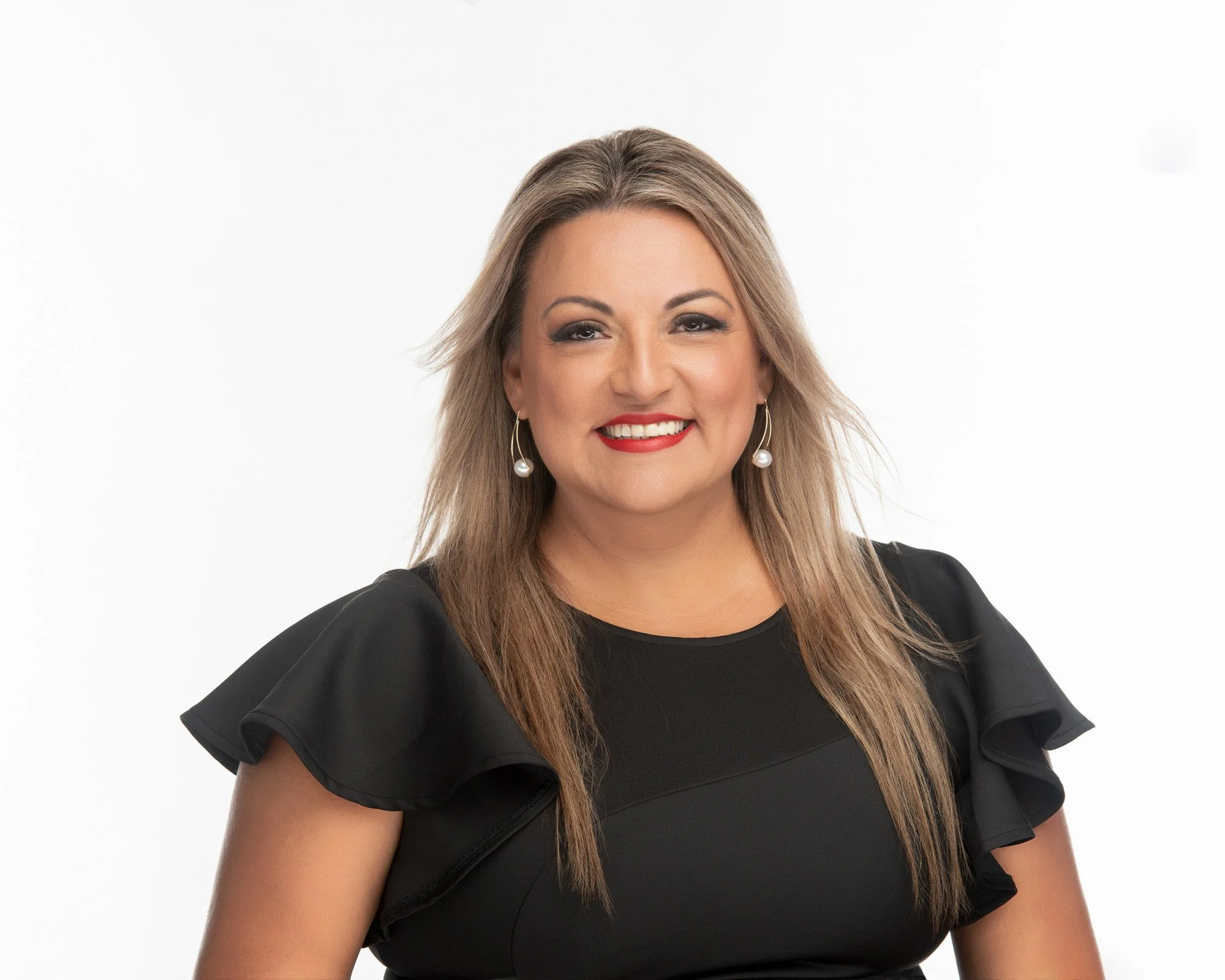 A woman with light brown hair, wearing red lipstick, pearl earrings, and a black dress with ruffled sleeves, smiling against a white background.