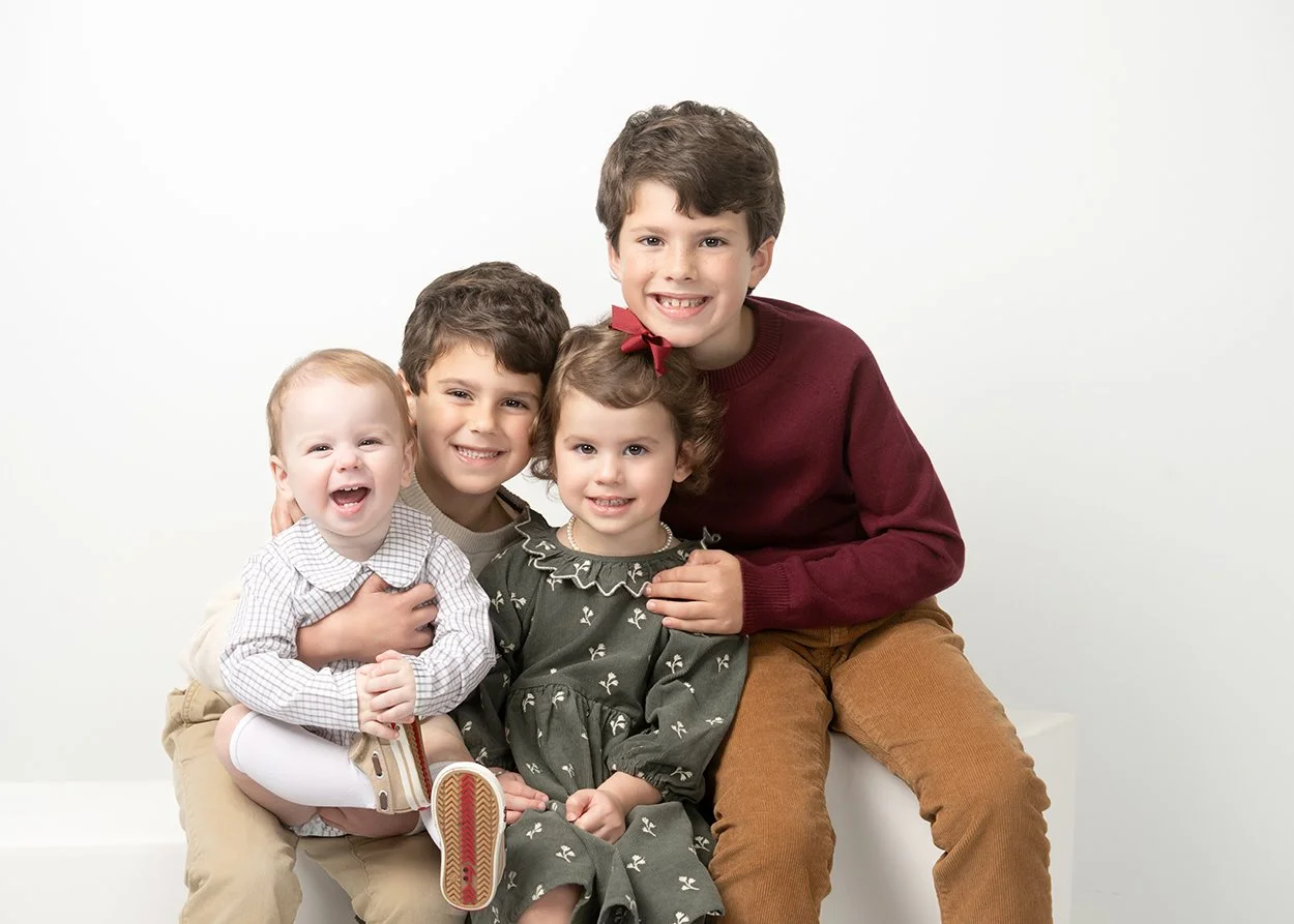 Four children sitting together, smiling, against a plain white background. The oldest boy is wearing a maroon sweater and brown pants. Next to him, a girl with a red bow in her hair is wearing a dark green dress with white floral patterns. The boy be