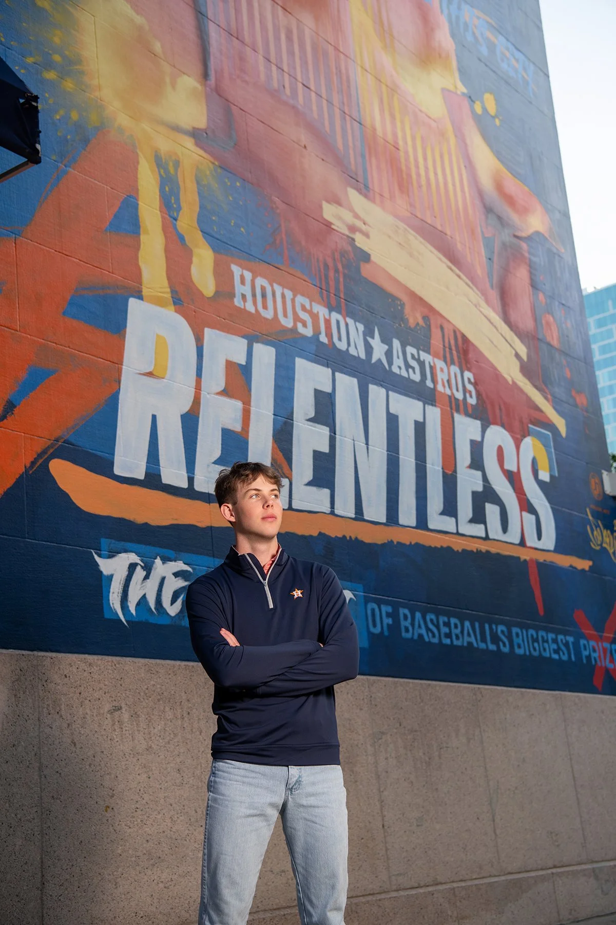 A young man with crossed arms, wearing a navy jacket and light jeans, standing in front of a colorful mural that celebrates baseball in Houston, Texas.