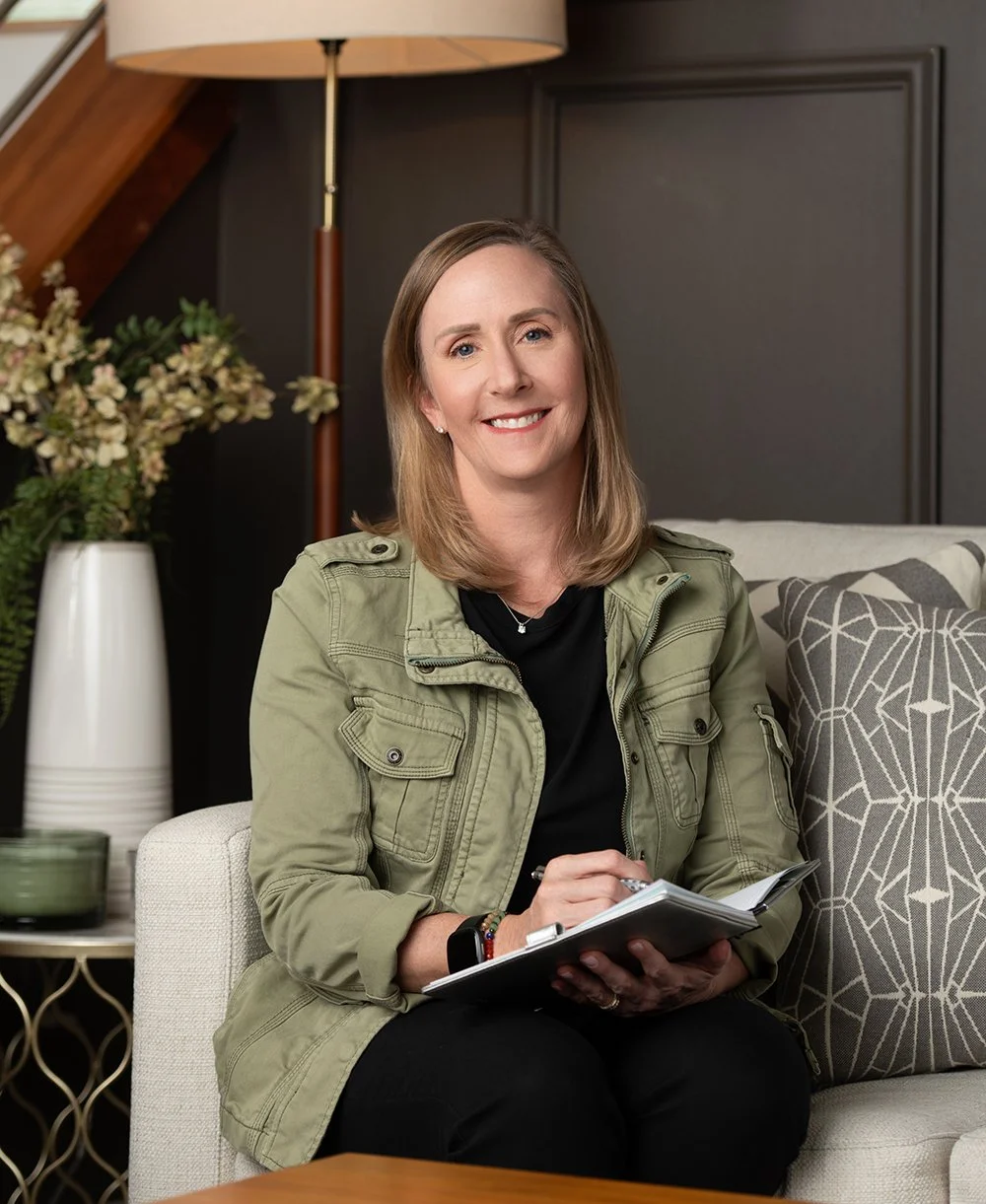 A woman sitting on a white sofa, smiling, holding a notepad and pen, in a cozy living room with a dark wall, a table lamp, a side table with plants and a patterned pillow.