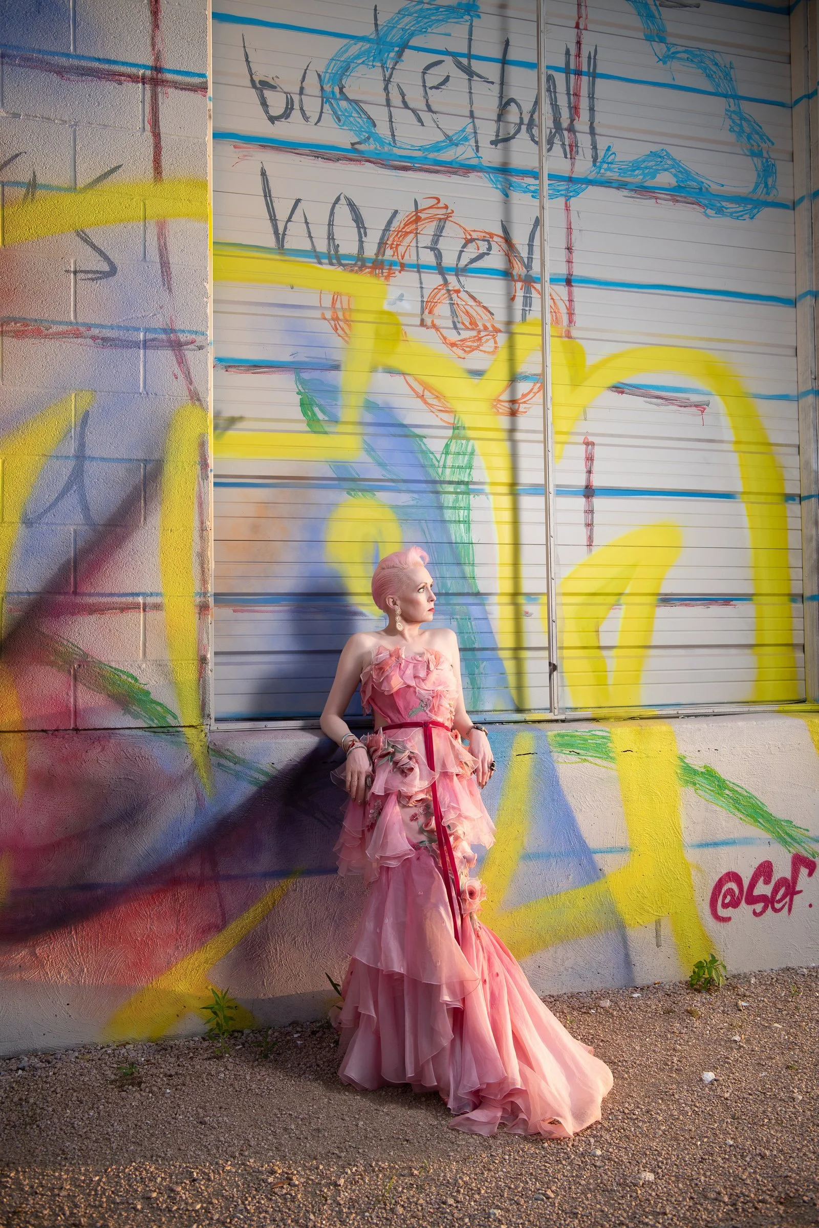 A woman with pink hair and a flowing pink gown standing against a graffiti-covered wall. The wall has colorful spray paint and the woman is looking upward.