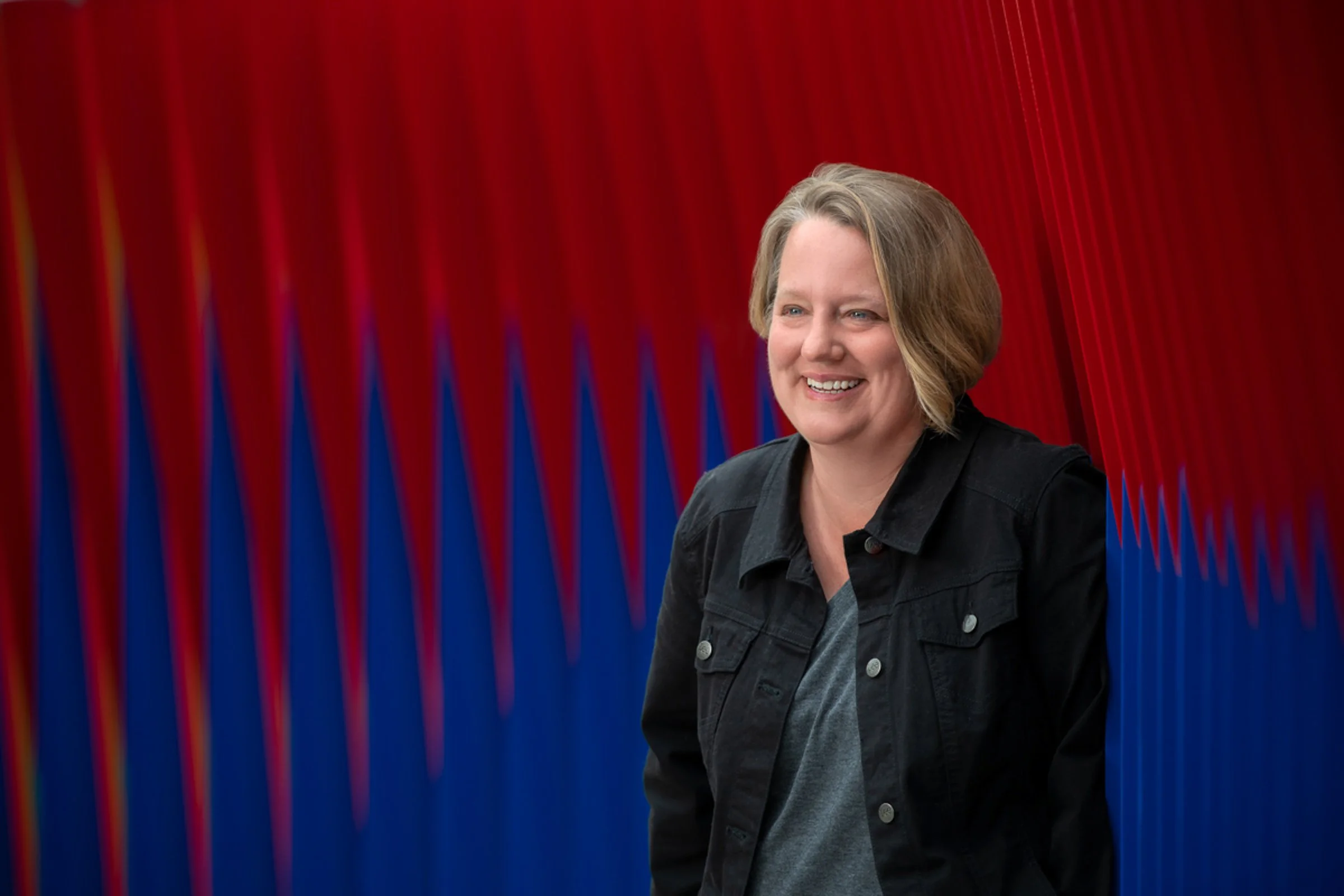 A woman with short blonde hair smiling, wearing a dark jacket, standing in front of a red, blue, and purple patterned background.