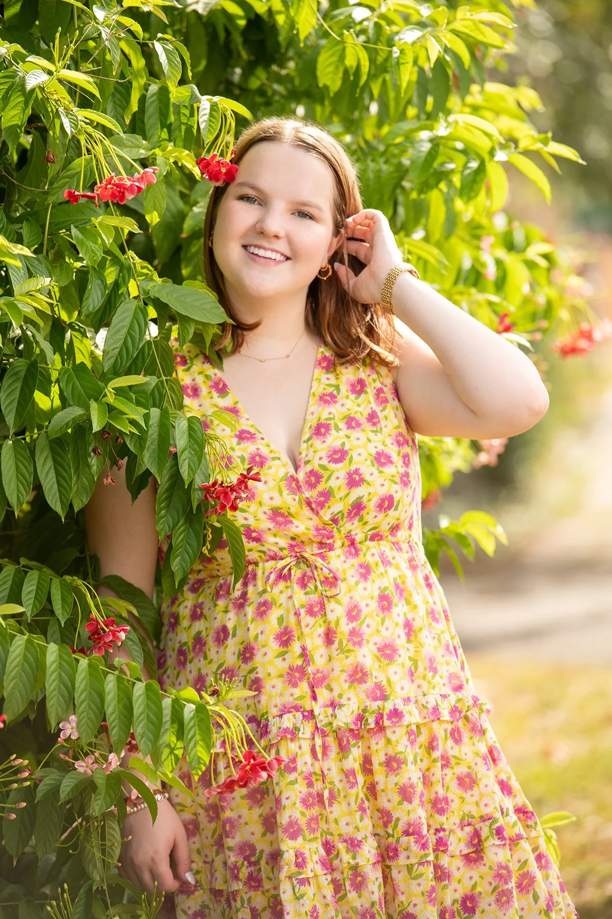 A woman in a yellow floral dress with pink flowers standing among green foliage and pink and red flowers, smiling at the camera.