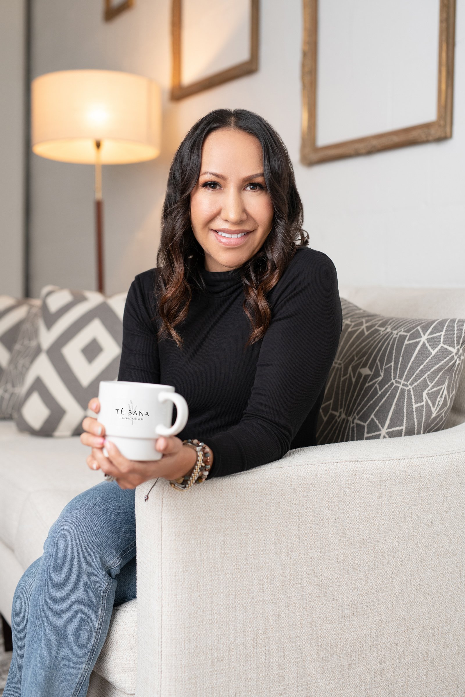 A woman with long dark hair, wearing a black long-sleeve top and blue jeans, sitting on a beige sofa, smiling, holding a white mug with "TÉ SANA" written on it, in a cozy living room with a lamp and decorative frames on the wall behind her.