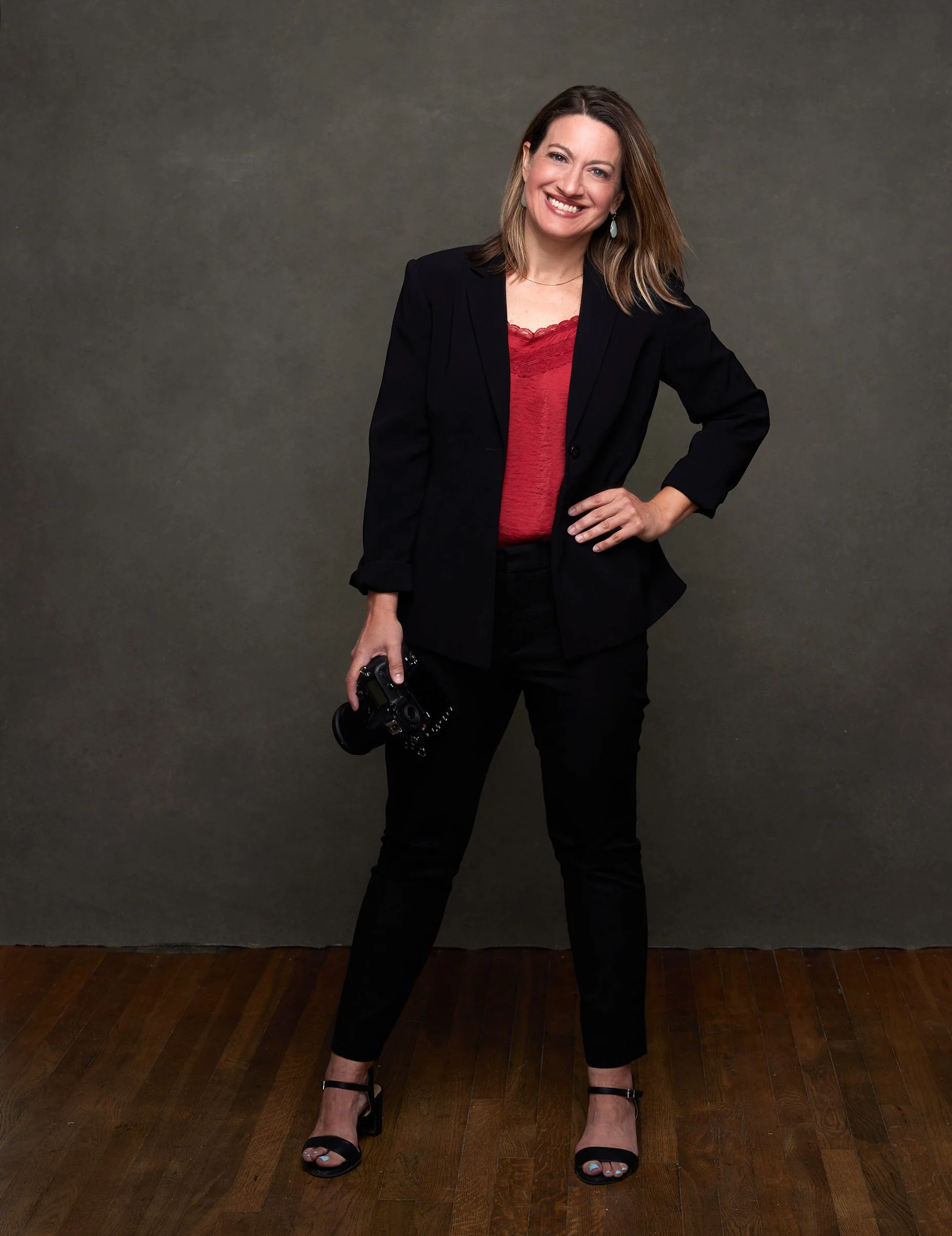 A woman in black pants, a black blazer, and a red top with lace trim standing on wooden floor with her hand on her hip, smiling at the camera, holding a camera in her other hand, against a plain gray wall.