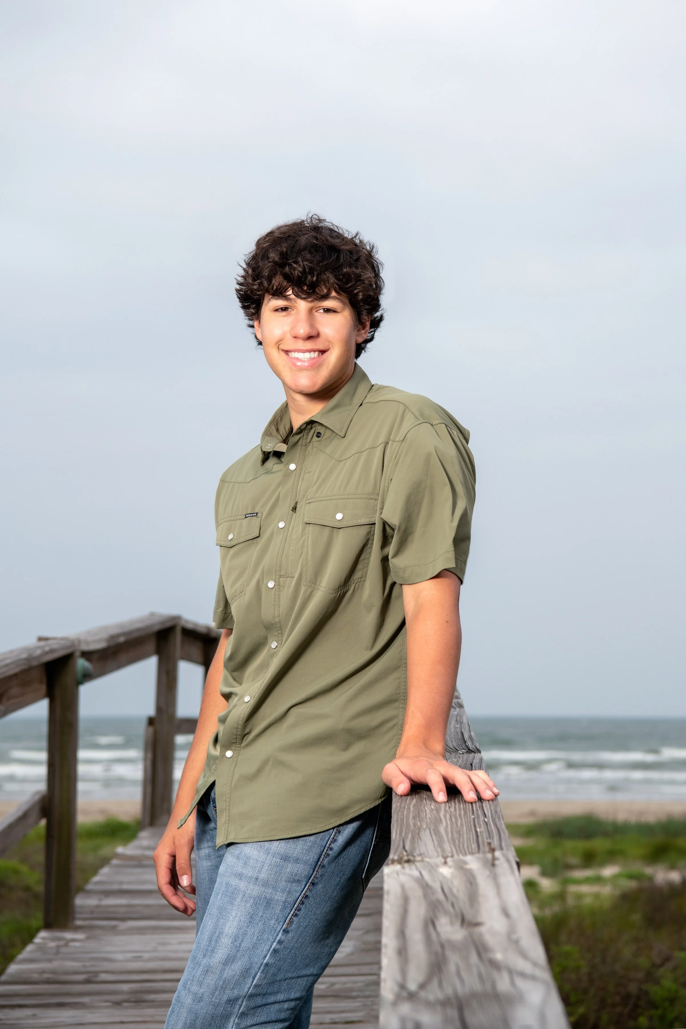 Young man with curly brown hair smiling, leaning on a weathered wooden railing on a beach boardwalk, wearing a green button-up shirt and jeans, with ocean waves and a cloudy sky in the background.