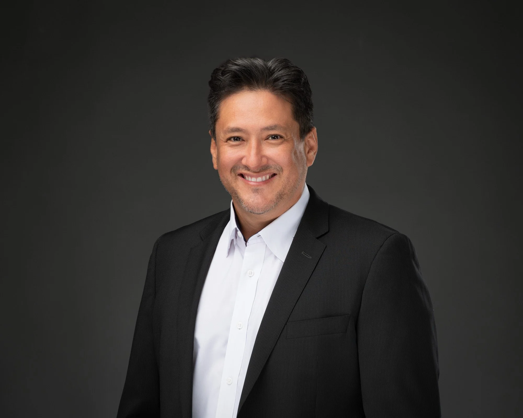 Portrait of a man with dark hair, styled back, smiling, wearing a black suit and white shirt, against a dark background.