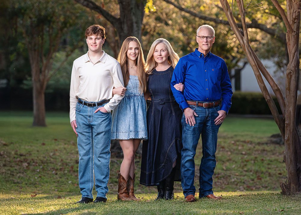 Family of four standing outdoors in a park during fall, smiling at the camera. The group includes a teenage boy, a teenage girl, a middle-aged woman, and an older man. They are dressed casually, with trees and sunlight in the background.