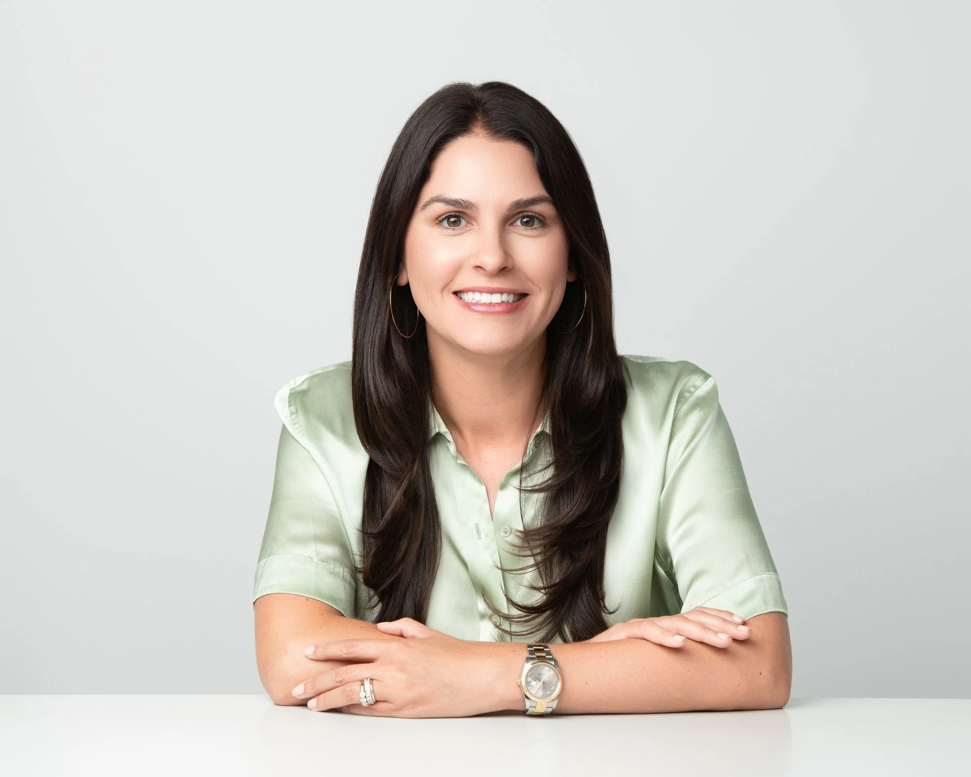 A woman with long dark hair smiling, wearing a light green blouse, gold hoop earrings, a watch, and rings, with her arms crossed on a white surface against a plain light gray background.