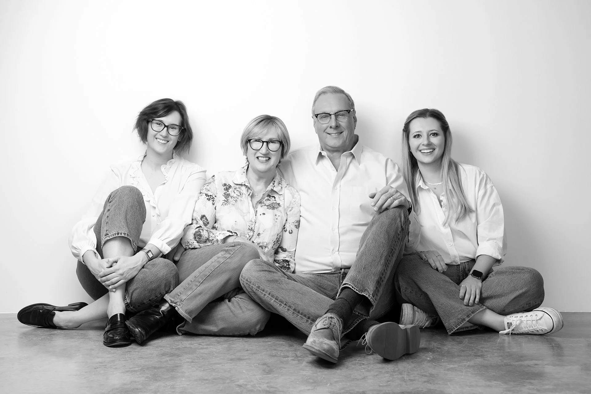 A family of four sitting and lying on the floor against a white wall, smiling at the camera. The group includes three women and one man, dressed casually.
