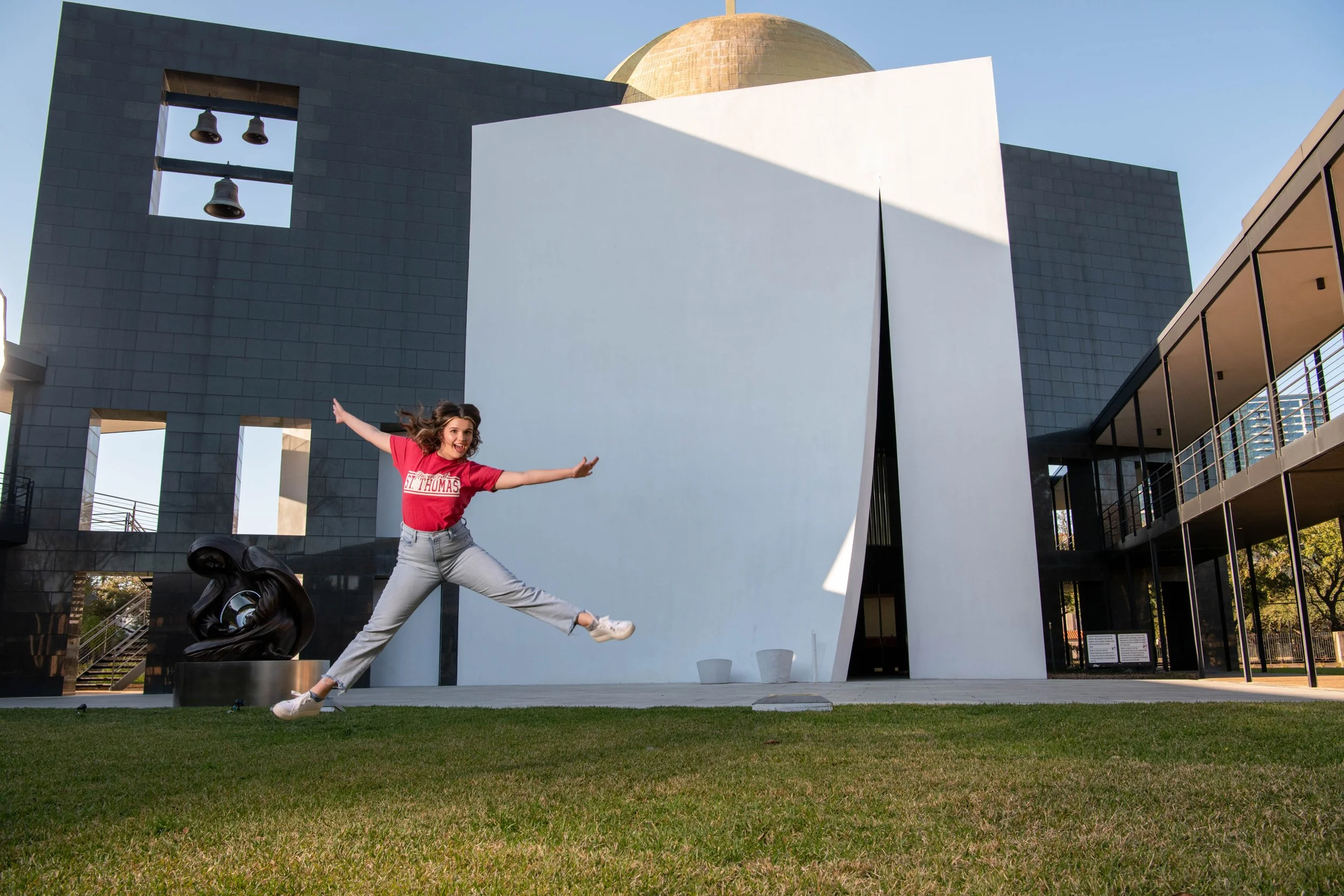 A woman in a red T-shirt and light gray jeans is jumping in front of a modern building with black, white, and beige architecture. She has her arms spread wide and looks excited, with a big smile on her face.