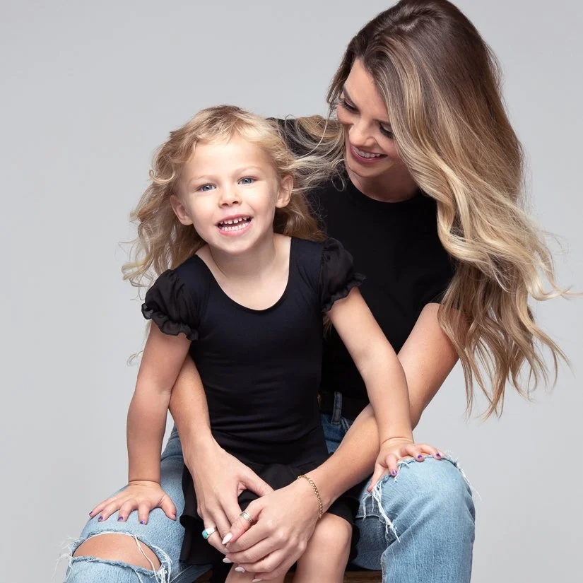 A young woman with long wavy blonde hair sitting with a happy blonde girl on her lap, both smiling and looking at the camera, against a plain gray background.